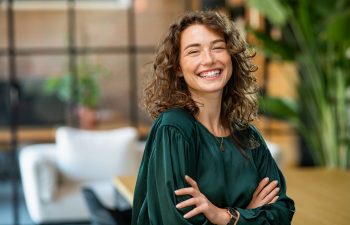 Smiling woman with curly hair wearing a green blouse, standing confidently in a modern wellness space, reflecting themes of skin health and rejuvenation at Skin & Body Medical Spa.