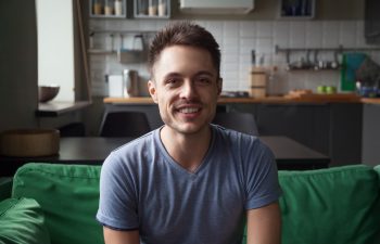 Smiling man in a casual blue shirt sitting on a green couch in a modern kitchen, representing a relaxed lifestyle related to wellness and effective weight loss solutions.